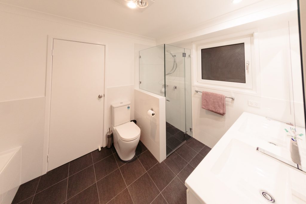 The fantastic new bathroom, a white wall-hung double-basin vanity and wall-mounted chrome taps are at the right of the photo. In the centre is a waist-high thin wall tiled in white, and that side of the shower screen starts at the top of the wall. Next to the wall sits a white ceramic toilet, and the toilet roll holder is a chrome metal one that's attached to the waist-height wall. The wall tiles are white and the floor tiles are a dark slate grey.