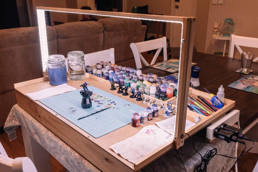 A photo of a DIY wooden "table" sitting on a dining table, with a square arch over the top of it with LED strip lights all along the inner surface of it. There's lots of Games Workshop paint pots and miniatures on it.