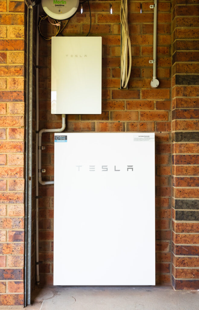A photo of a white Tesla Powerwall 2 battery and Backup Gateway mounted against a red brick wall inside our garage.