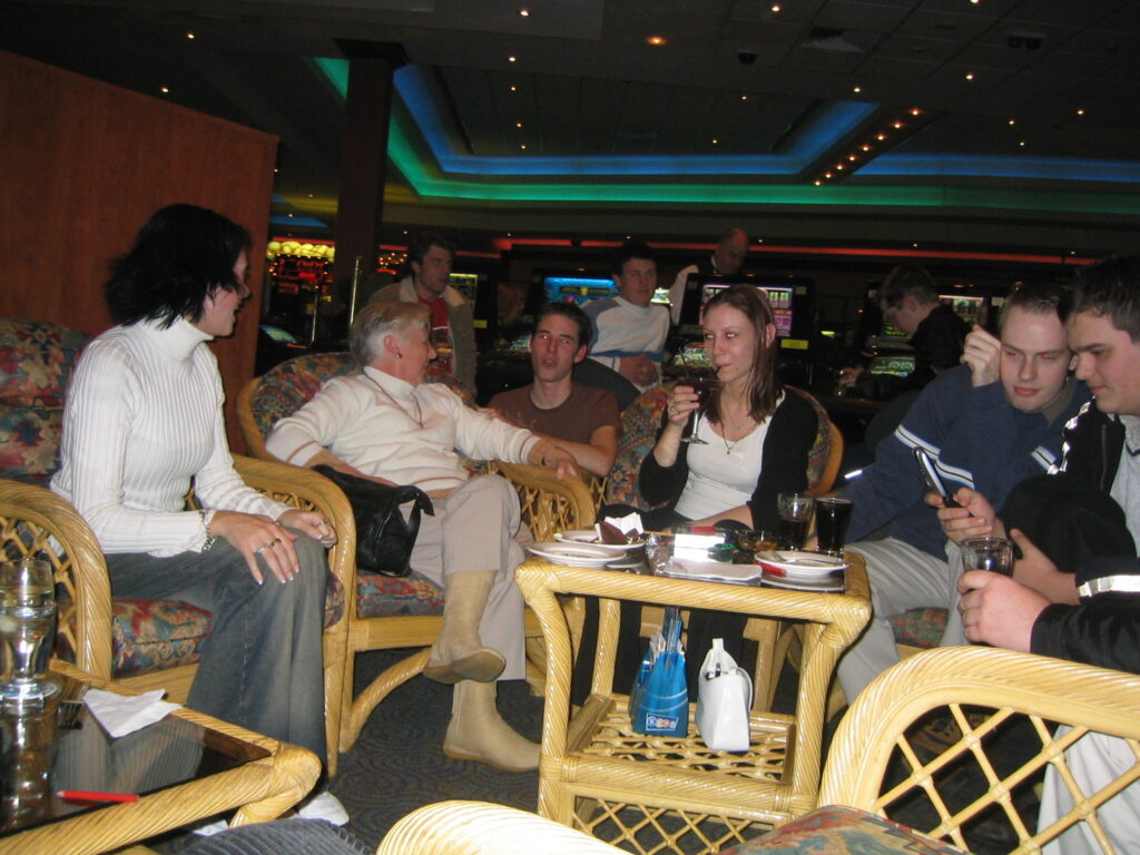 A group of people sitting around in very daggy-looking wicker chairs with a bunch of pokies in the background. The camera flash is on so the lighting is terrible.