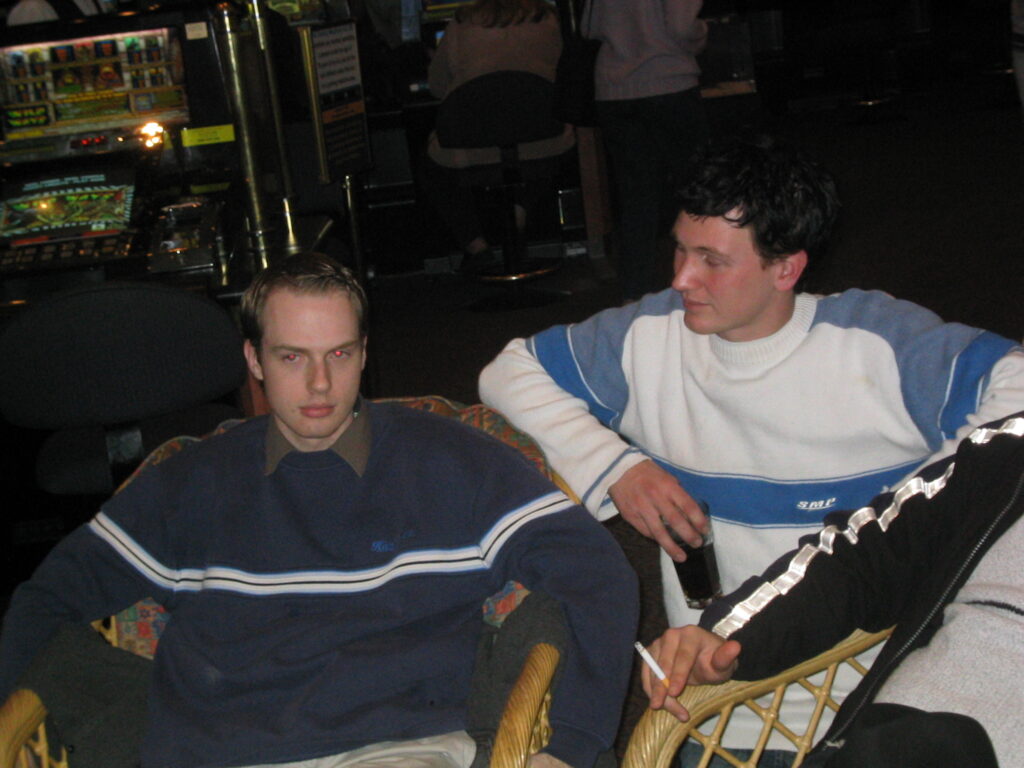 A photo of an extremely young me, a beardless white man with short but gelled-up brown hair sitting in one of the daggy wicker chairs. Another guy with slightly longer brown and scruffy hair is crouched next to me like we were in discussion about something.